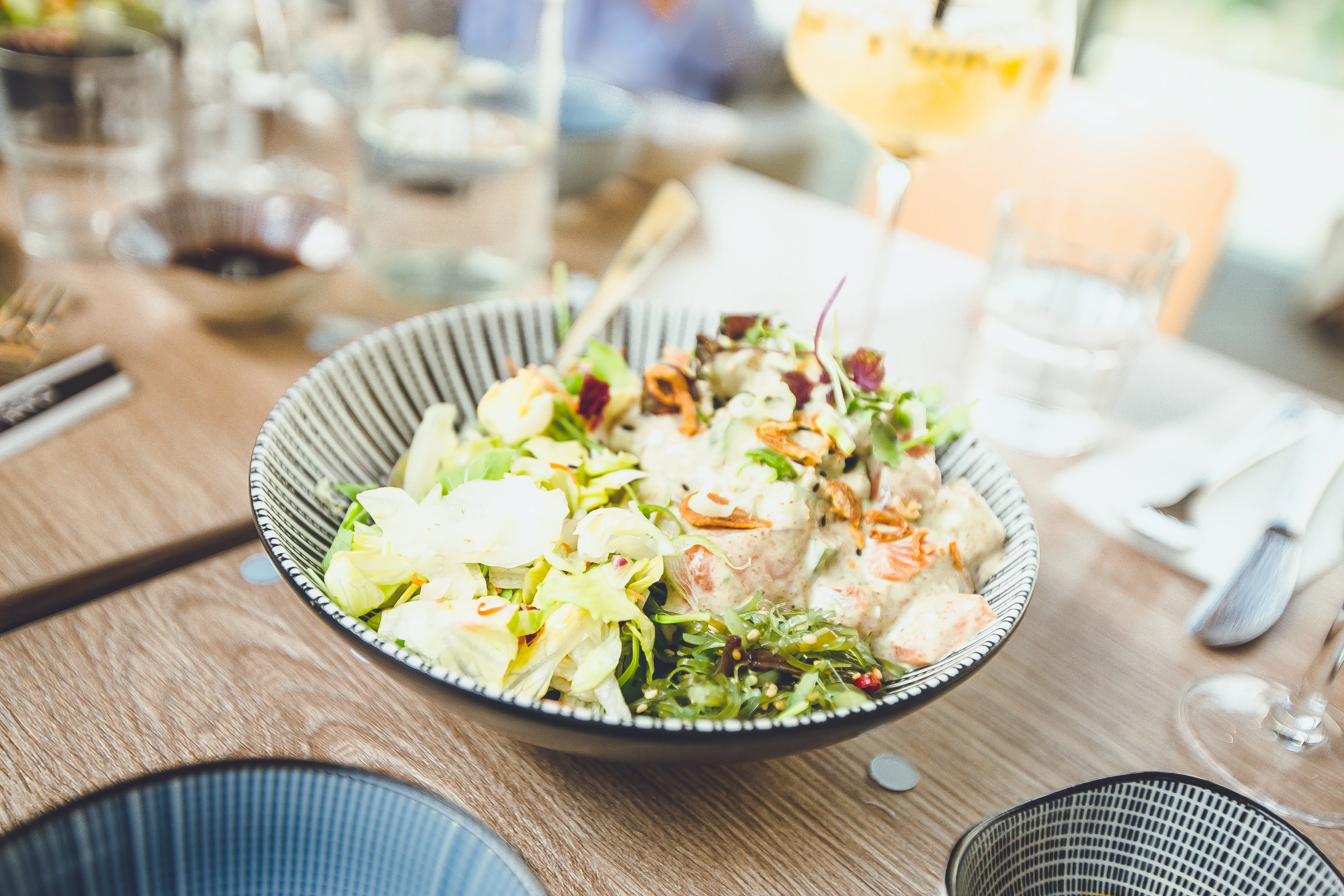 Frische Poke Bowl mit Salat und Dressing in einer gemusterten Schüssel auf einem Holztisch in einem Restaurant
